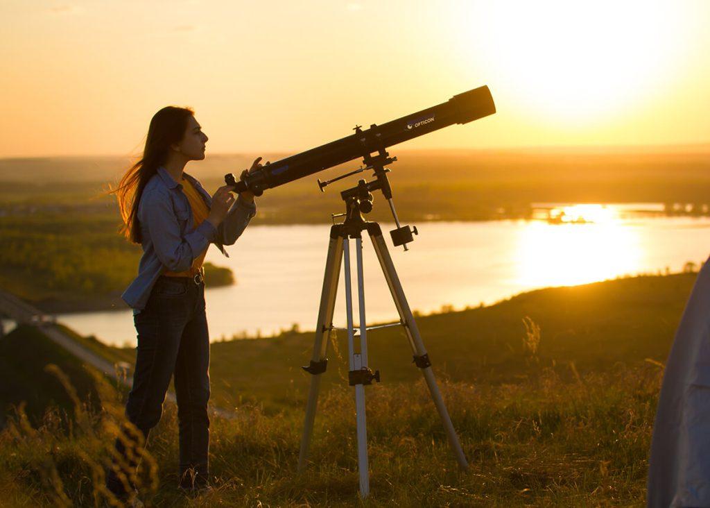 kobieta z teleskopem na łące nad jeziorem o zachodzie słońca