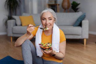 Sports and nutrition concept. Happy senior femael leaning eating fresh vegetable salad, sitting on yoga mat at home