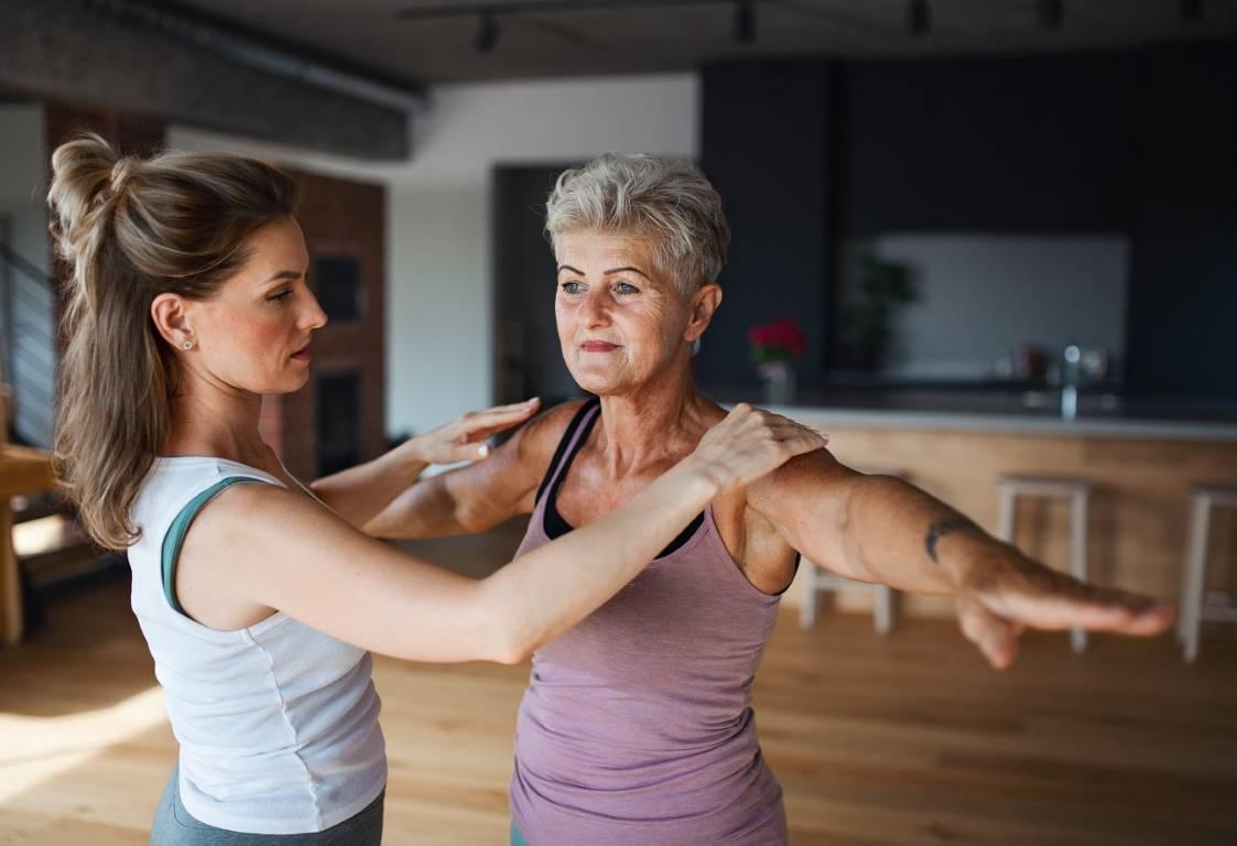 Active senior woman in sportsclothes exercising with female physioterapist indoors at home.